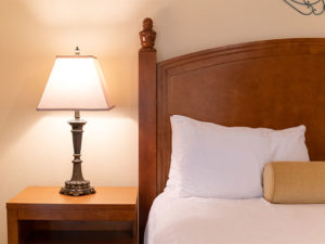Close up of bedside table with table lamp, wooden bed headboard, white pillow and sheets with tan accent pillow