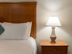 guest room up close photo of bedside with light brown headboard and nightstand, white pillows, white textured bed linens and white table lamp
