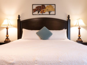 Master bedroom bed straight on with dark brown headboard and nightstands, table top lamps, white pillows and white textured bed linen