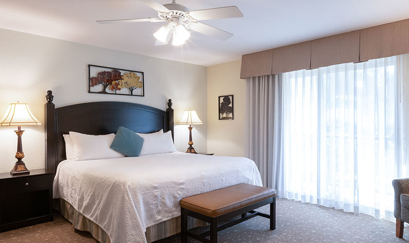 Master bedroom with light tan patterned carpet, bed with dark brown headboard, nightstands and bench. White ceiling fan and floor to ceiling window